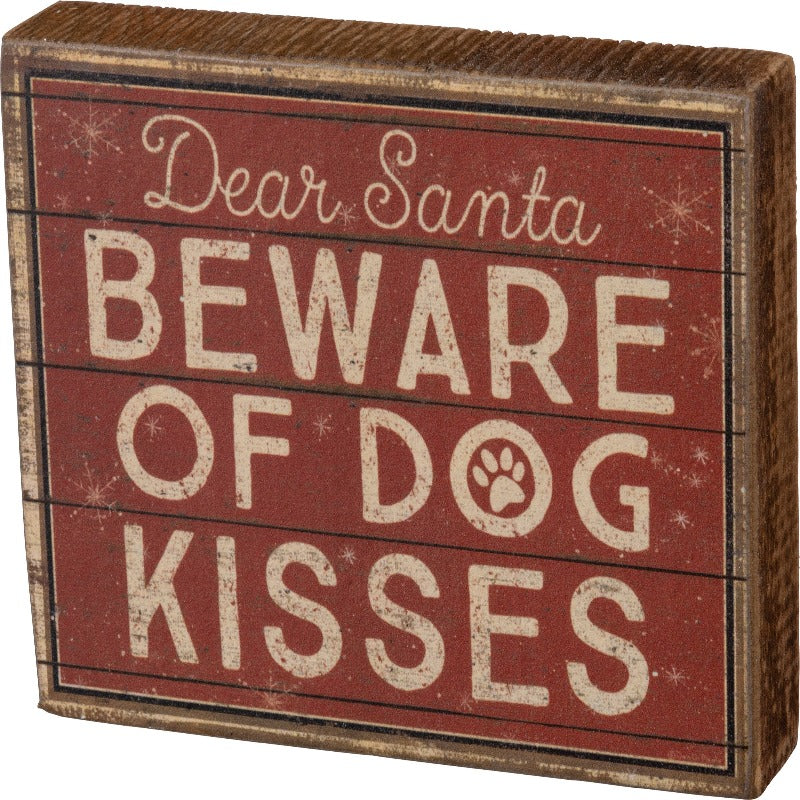 This rustic-inspired red block sign featues a "Dear Santa Beware Of Dog Kisses" sentiment, paw print and snowflake designs, and slat wood background print.