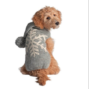 A small fluffy dog wearing a gray knit pet sweater with a white snowflake pattern and a pom-pom hood, shown against a plain white background.