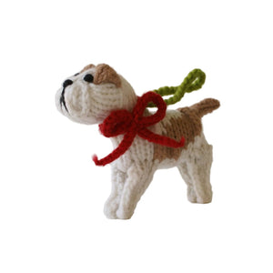 A small crocheted dog ornament or figurine in cream and tan, wearing a red bow and green loop, photographed against a plain white studio background.