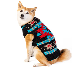 A Shiba Inu dog wearing a black knit sweater with a colorful geometric pattern, photographed against a solid white background.