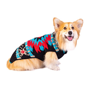 A fluffy tan and white Corgi wearing a colorful knit dog sweater with a black base and red, blue, and beige geometric patterns, photographed against a solid white background.
