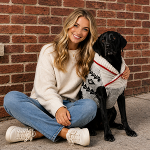 Woman sitting on the ground with a black dog wearing a sweater against a brick wall.