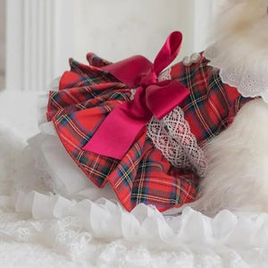 A small white fluffy dog wearing a red and green tartan plaid pet dress with a large hot pink satin bow and white lace trim, resting on a white ruffled blanket.