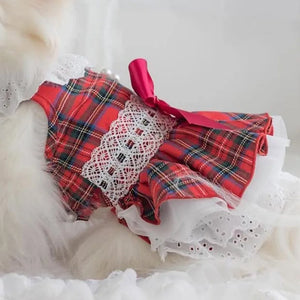 A red tartan plaid pet dress with white lace trim and a pink ribbon bow, shown on a white fluffy dog.