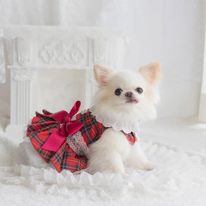 A small white fluffy dog wearing a red and green tartan plaid dress with a large red bow and white lace collar, sitting on a white ruffled surface in front of a white decorative fireplace.