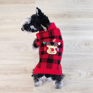 Dog wearing a red and black checkered sweater with a reindeer design on a wooden floor.