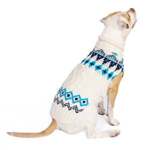 A dog wearing a white knit sweater with navy and teal geometric patterns, photographed against a plain white background.
