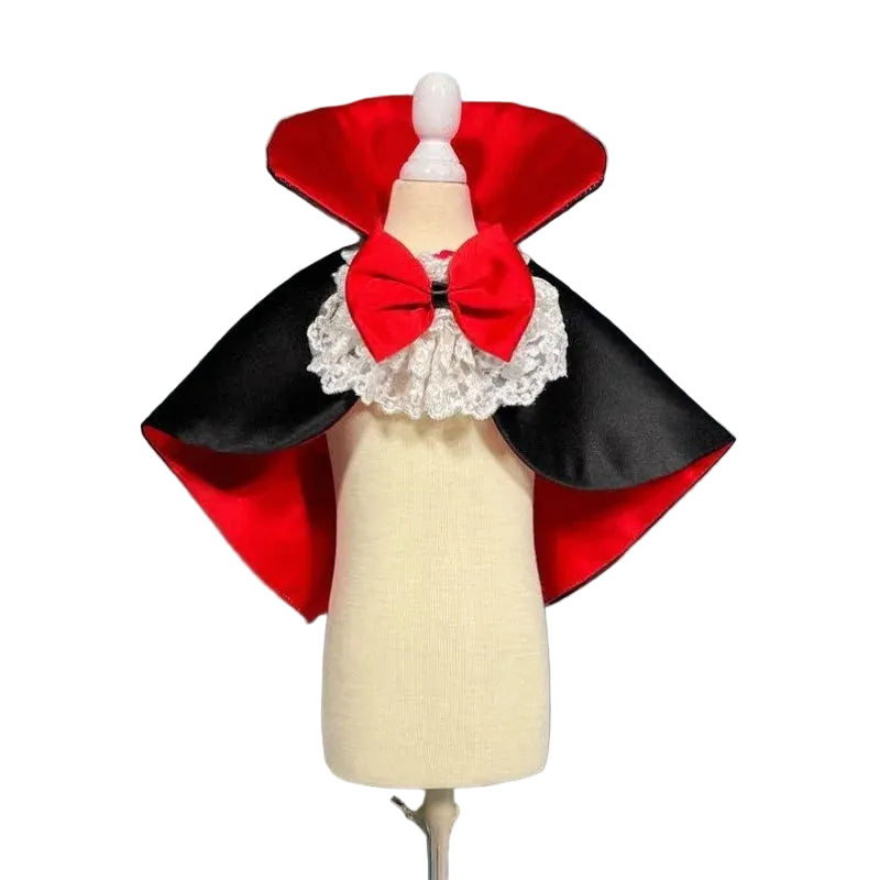 A small white dog wearing a vampire costume with a black cape, red collar, and red bow, sitting on a white surface against a blue-gray paneled wall.