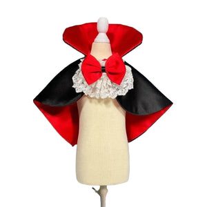 A vampire-style dog costume cape displayed on a cream dress form mannequin. The cape has a bright red inner lining, black satin outer wings, and a large red bow with a white lace ruffle collar at the neck.