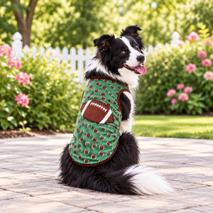 A black and white Border Collie dog sitting outdoors on a paved walkway, wearing a green dog coat with a brown football pattern and a football patch on the back.