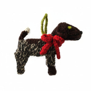 A handmade knitted dog ornament in dark brown, cream, and black yarn, featuring a bright red knitted bow around its neck, a green hanging loop, and small green and white eyes, set against a plain white background.