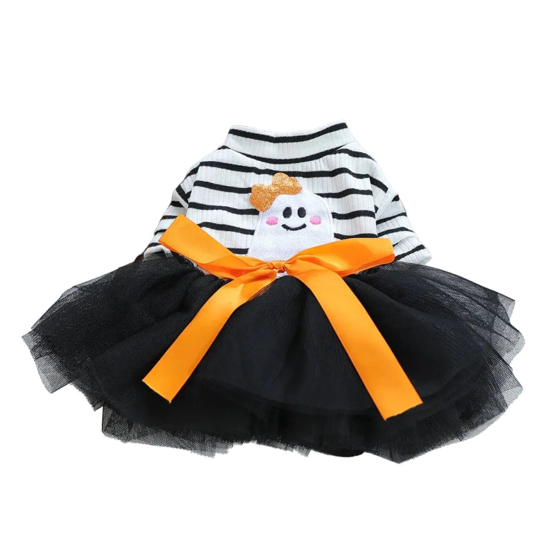 A small Yorkshire Terrier sits on a white window sill wearing a Halloween-themed pet dress. The dress features a black and white horizontally striped knit top with a white ghost applique, a black tulle skirt, and a large orange satin bow.