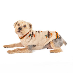 A small tan and gray dog lying on a white background, wearing a beige knit pet sweater with orange, brown, and green decorative motifs.