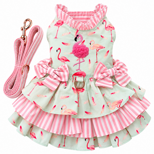 A pink and mint green dog dress with a flamingo print, featuring a ruffled collar, tiered skirt, and two large bows, shown with a matching pink leash on a white background.