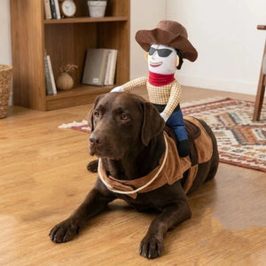 A brown Labrador retriever lies on a wooden floor wearing a cowboy costume, with a cowboy doll sitting on its back. The scene is set in a cozy indoor room with a bookshelf and patterned rug in the background.