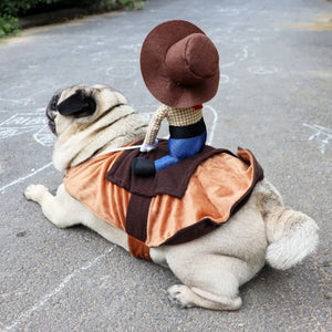 A pug dog lying on asphalt wearing a brown and orange costume vest, with a cowboy doll sitting on its back. The dog is photographed outdoors on a paved surface with chalk drawings in the background.