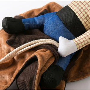 A close-up of a plush cowboy doll wearing blue jeans, brown boots, a beige and red plaid shirt, and a brown hat, resting on a brown and dark brown fleece blanket with a cream rope.