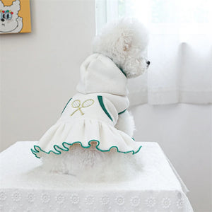 Small white dog wearing a white dress with green trim on a light-colored table.
